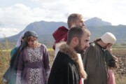 Personaggi in costume camminano fra i campi arati con le montagne sullo sfondo
