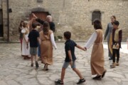 Bambini in tunica ballano in girotondo guidati da un adulto in un cortile